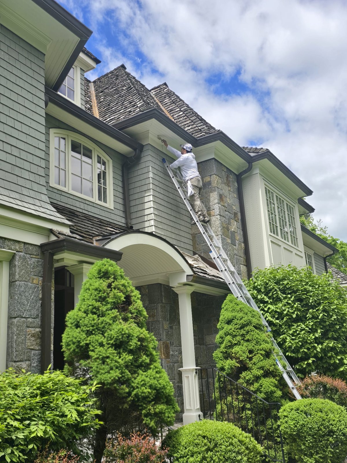 L&J Painting Group craftsman on ladder painting exterior trim of a grand shingle-style home
