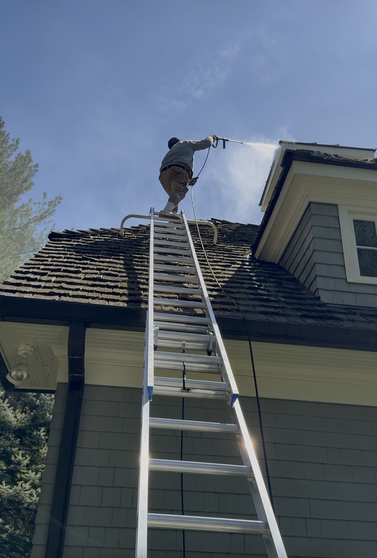Technician on an extension ladder power washing the gutter and roofline of a home against a clear blue sky