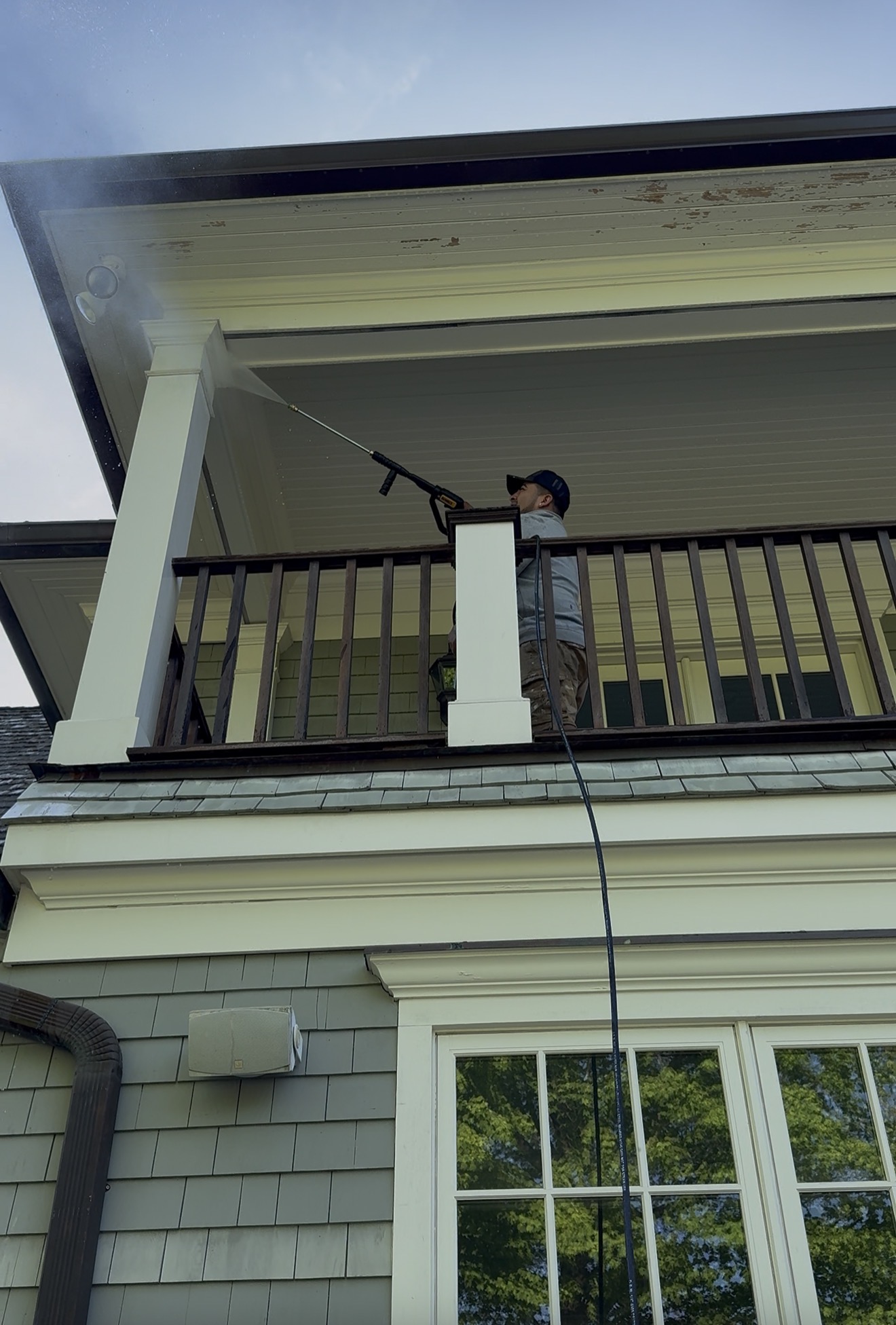 Technician power washing the soffit and roofline from a second-floor porch on a shingle-style home