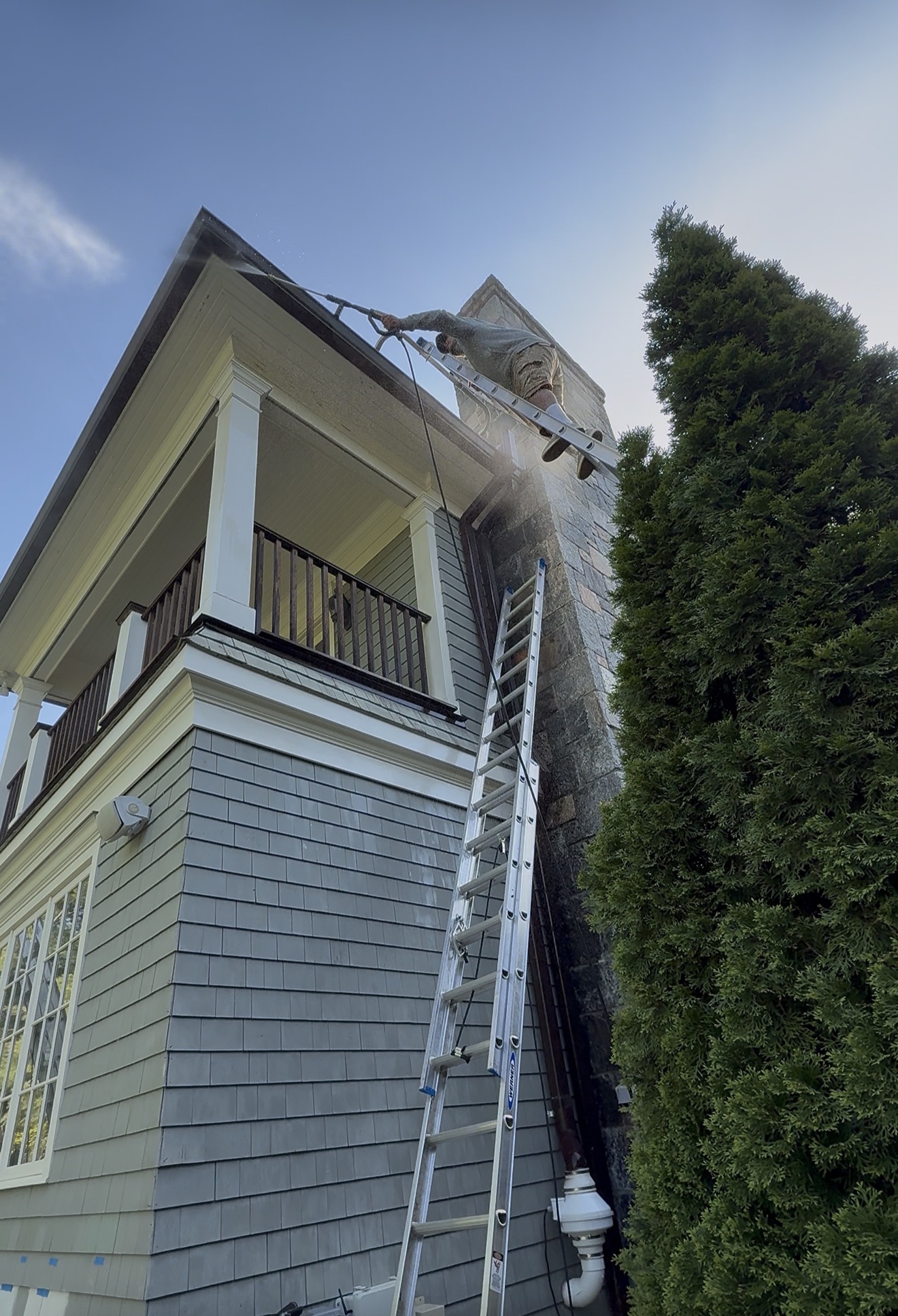 Power washing spray reaching the upper eave of a tall coastal-style home with a rooftop lookout tower