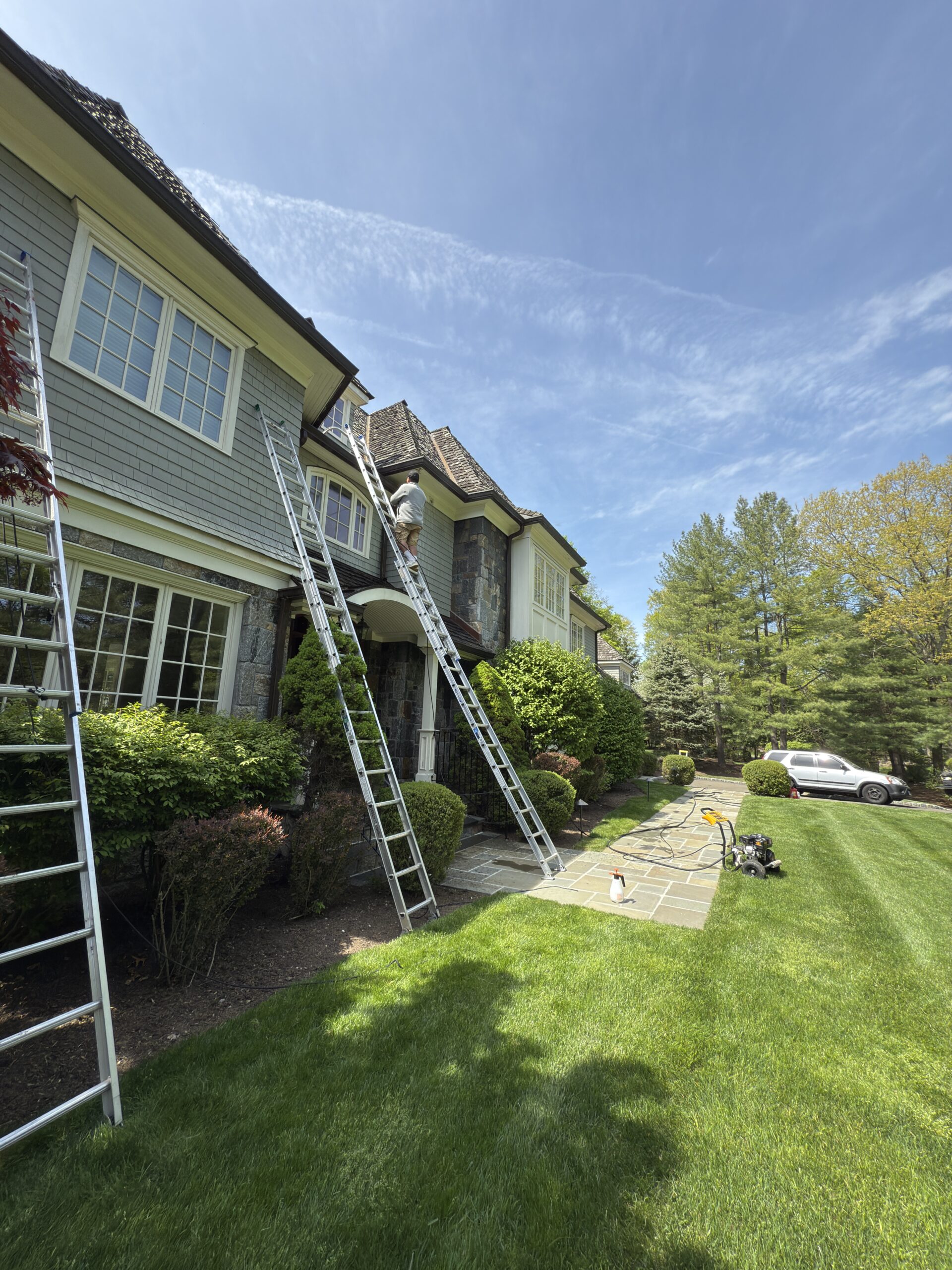 Large shingle-style home prepared for power washing with extension ladders positioned at the roofline and entry